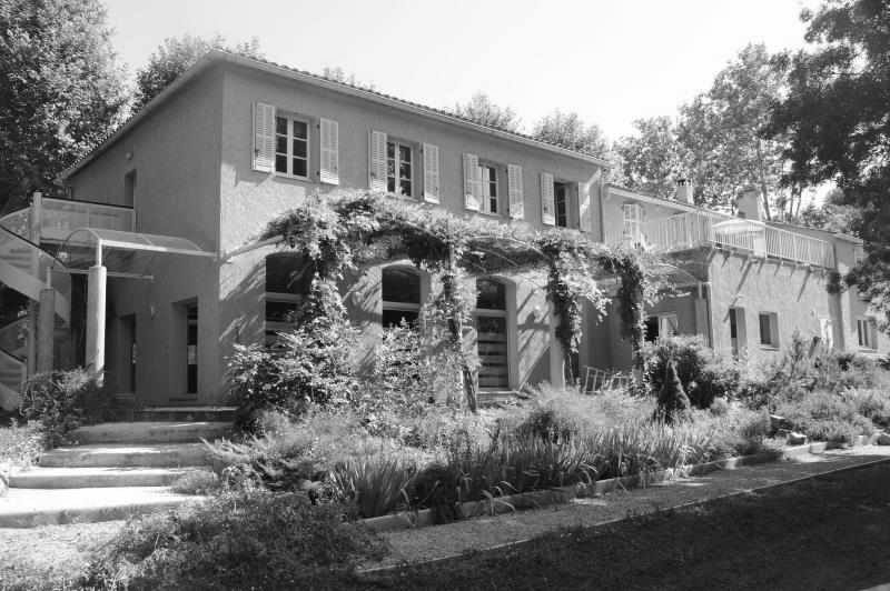 Black-and-white photo of the Shumeikan at Bras: a two-storey Provençal building with shuttered windows, a vine-covered pergola along the ground floor, stone steps and a planted garden in front, with tall trees behind.