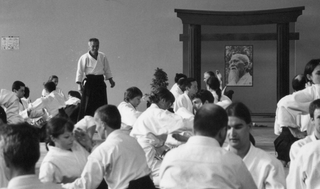 Black-and-white photo: Tamura Shihan, in white keikogi and dark hakama, stands watching rows of students practising in pairs on the mat at Lesneven; a portrait of O-Sensei hangs on the dojo wall behind them.
